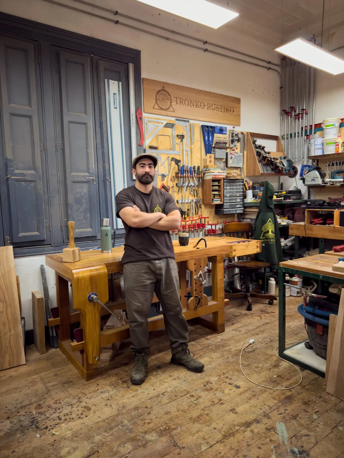 Matías Astudillo de brazos cruzados en el taller, junto al mesón de carpintero Roubo y el letrero de Tronko Rústiko.